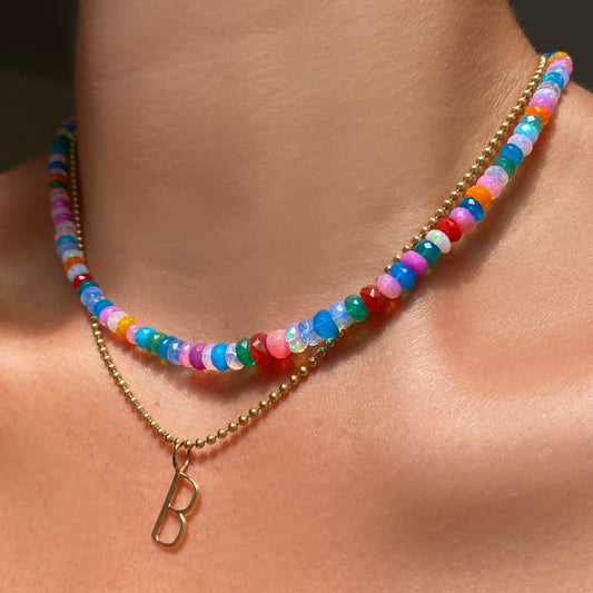 Shimmering beaded necklace made of faceted opals in shades of fiery blues, yellows, teal, and purple on a gold linking oval clasp. Styled on a neck layered with bead chain necklace and letter B charm.