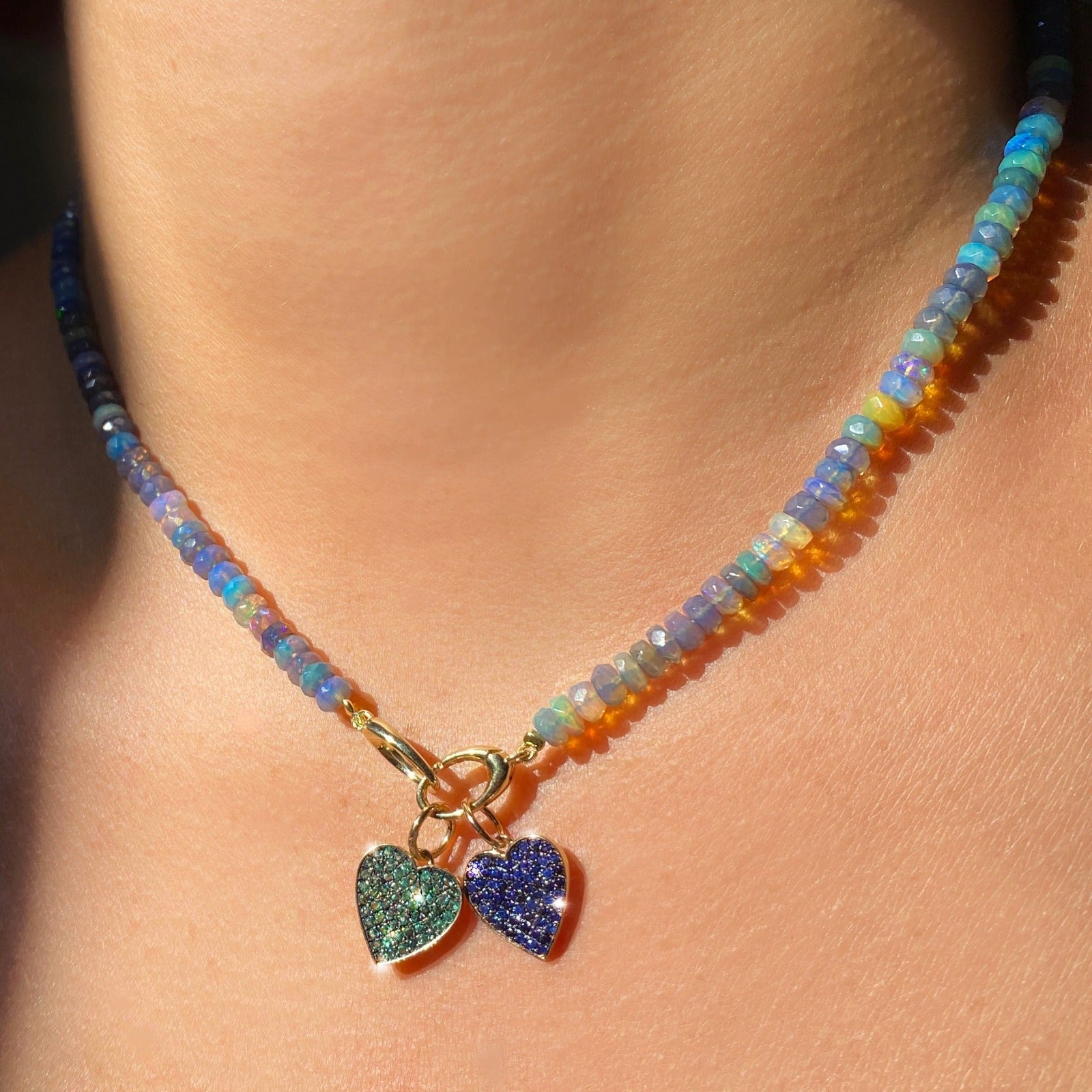 Shimmering beaded necklace made of faceted opals in shades of light blue, light purple, yellow, white, and clear on a gold linking ovals clasp. Styled on a neck with two medium pave heart charms.