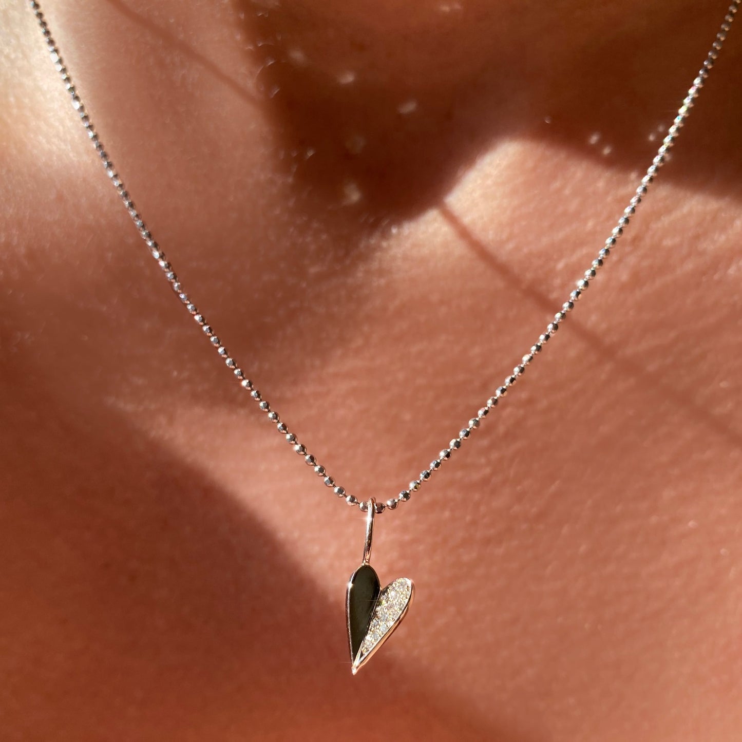 14k white gold small folded heart charm with pave diamonds on the right side. Styled on a neck hanging from a diamond cut bead chain necklace.