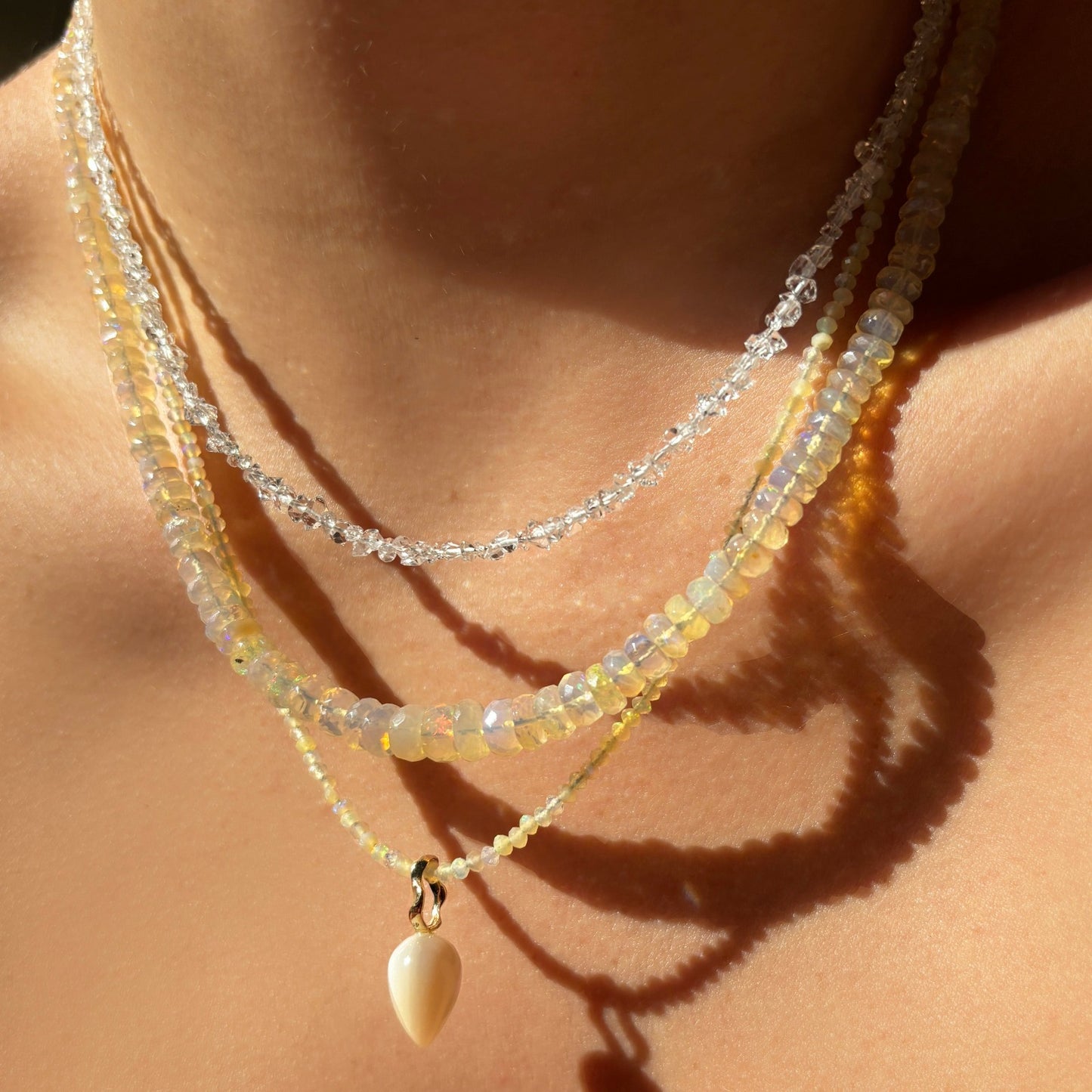 Shimmering beaded necklace made of faceted opals in shades of clear and nude opals on a gold linking ovals clasp. Styled on a neck layered with an acorn drop charm.