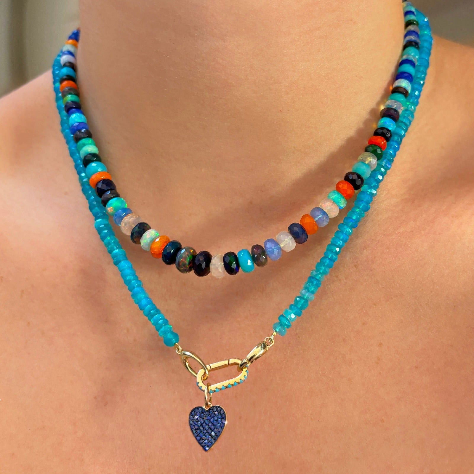 Shimmering beaded necklace made of faceted opals in shades of bright light blue on a gold linking ovals clasp. Styled on a neck layered with a small turquoise charm and medium pave heart charm.