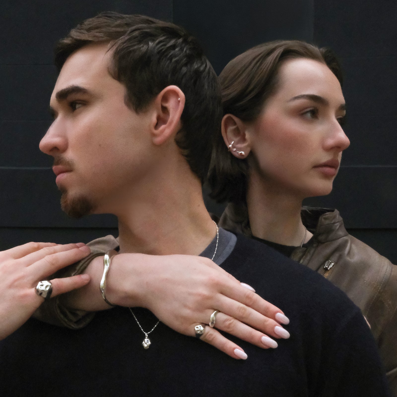 Two people standing close together with one person's hand on the other's shoulder, both wearing AMANDA PEARL molten sterling silver jewelry, against a dark background.