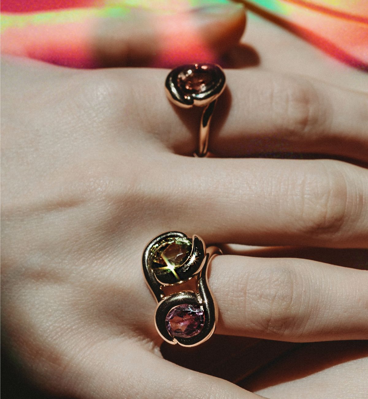Three gold rings are worn by a model in a close-up of her hand. These Molten Knot rings feature one of a kind stones, amethyst, green tourmaline, and green brown tourmalines. The stones shine as do the 14K recycled gold bezel setting.