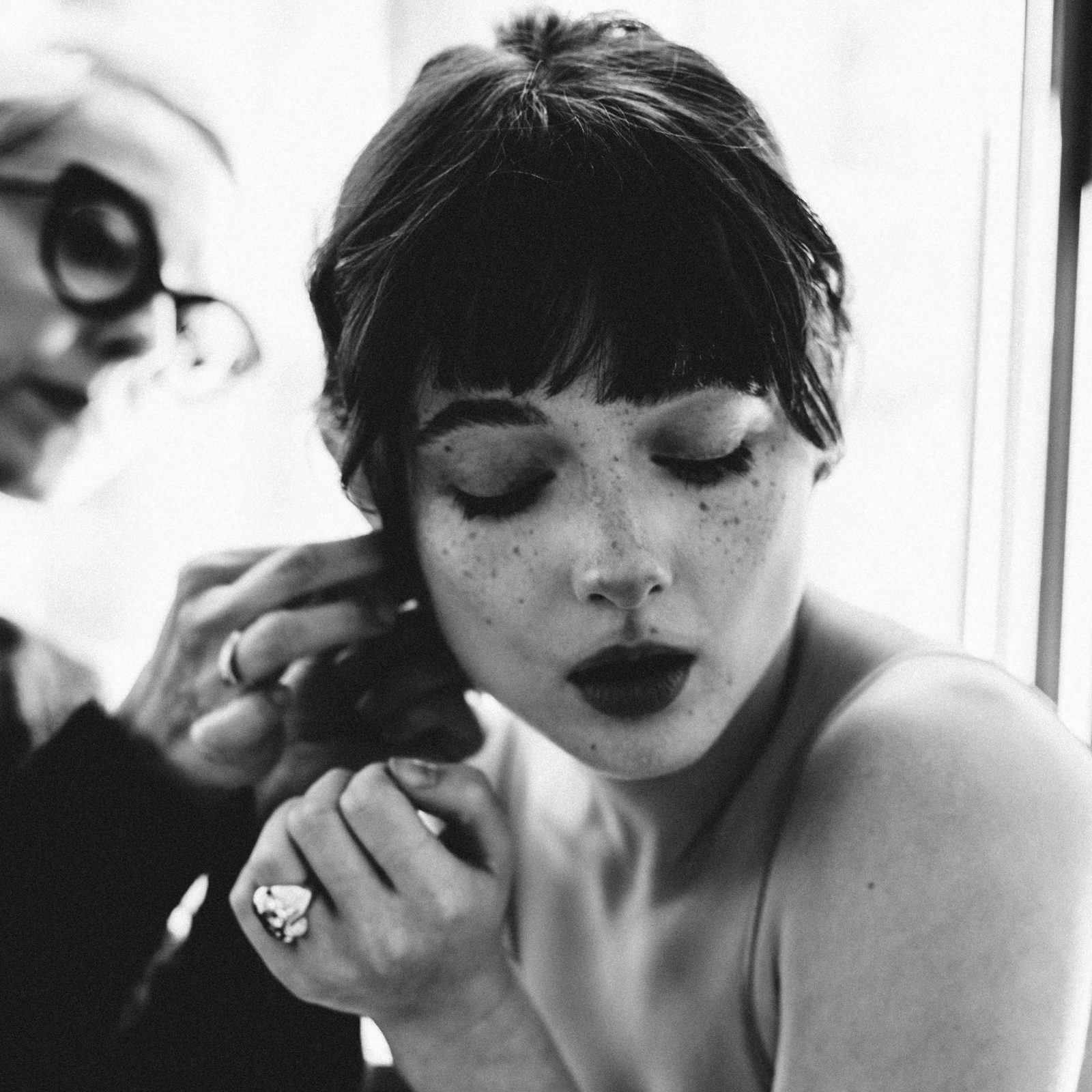A Black and white close-up of a woman having her AMANDA PEARL adjusted by Amanda Brotman, with a bright window in the background.