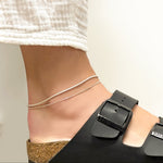 Sterling Silver foxtail chain anklet layered over a diamond cut bead chain anklet on a model's ankle, wearing white cotton gauze pants and black Birkenstock Arizona sandals.