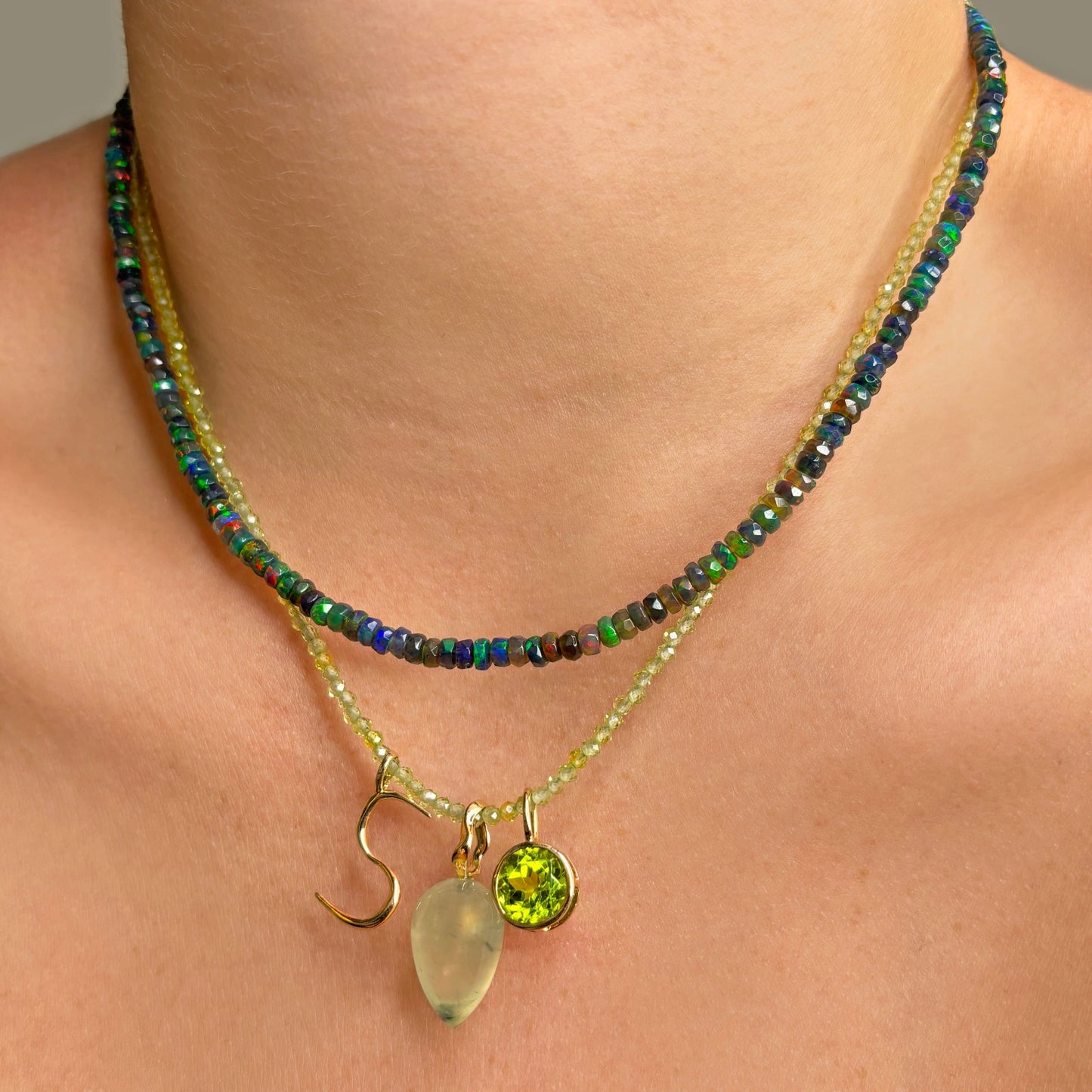 Shimmering beaded necklace made of 2mm faceted opals in shades of light green on a gold linking lobster clasp. Styled on a neck layered with an S letter charm, acorn drop charm, and round solitaire charm.