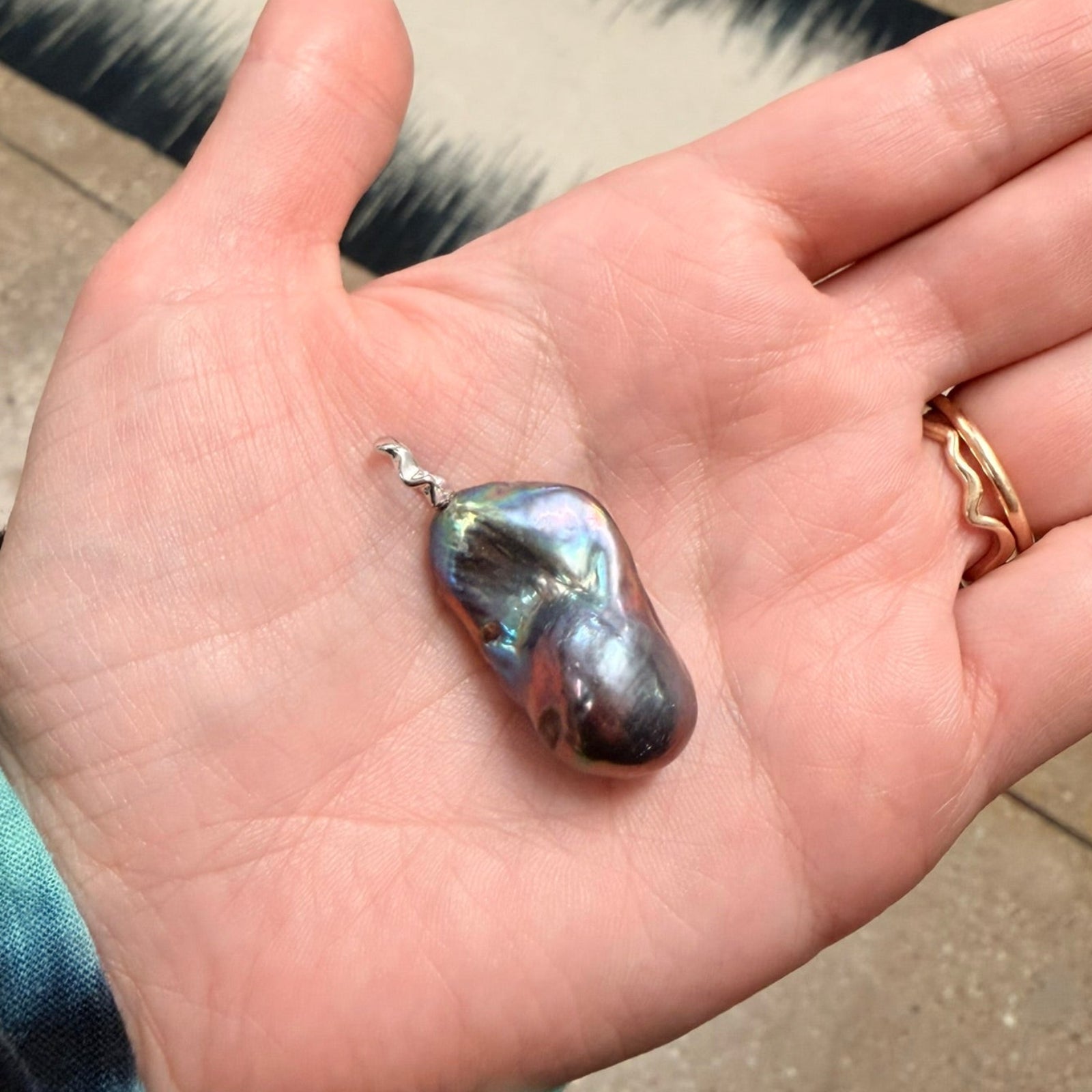Storm blue / purple / peacock Baroque Pearl Charm with recycled 14k white gold ripple bale in a person's hand.