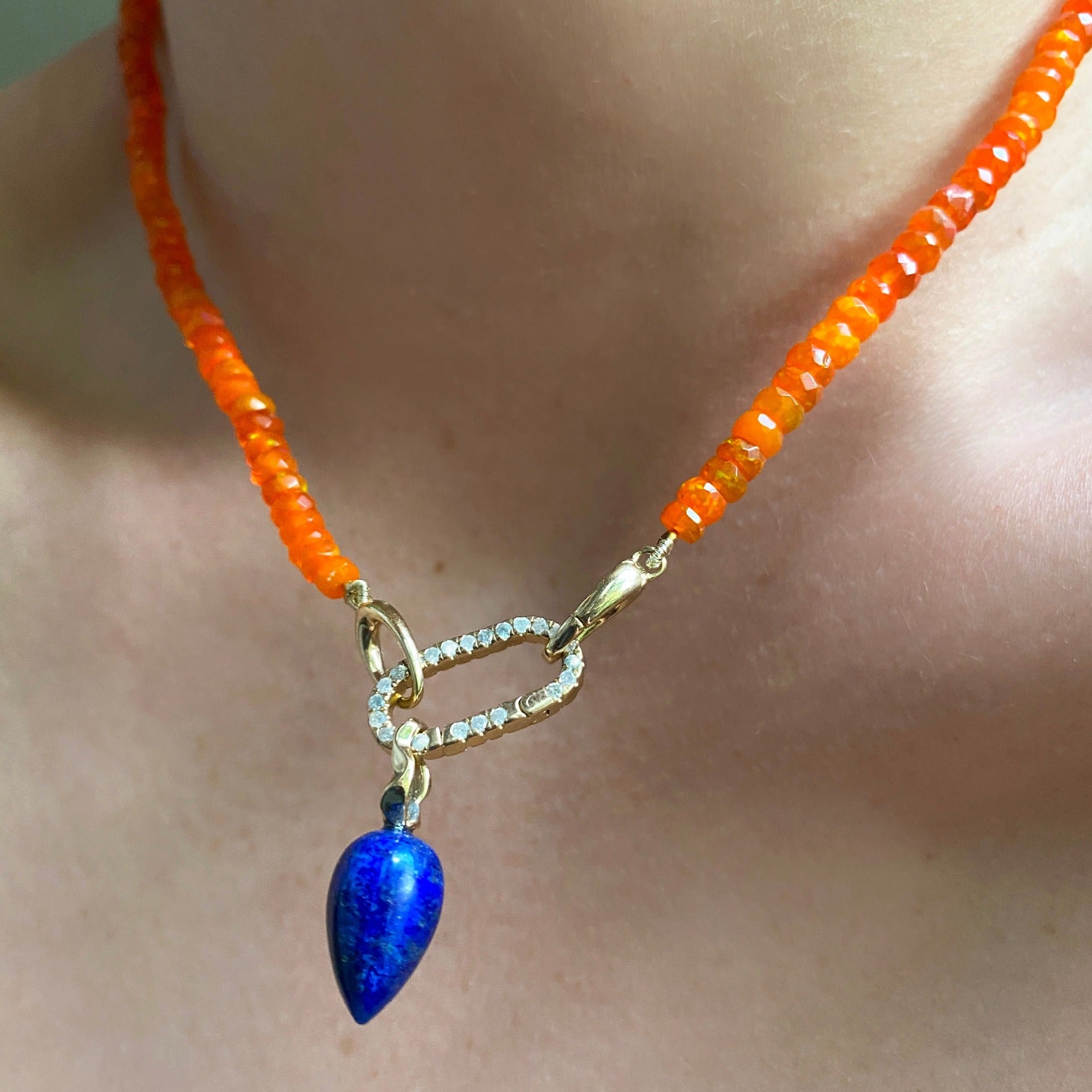 Shimmering beaded necklace made of faceted opals in shades of orange on a gold linking ovals clasp. Styled on a neck layered with a medium pave face charm lock and a lapis acorn drop charm