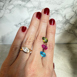 A close-up of a hand with dark red nail polish wearing a 14K yellow gold pear-cut diamond ring, with three alternative colorful gemstones set on her hand. They are peridot, pink topaz, and blue topaz.