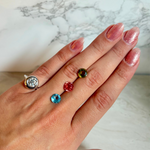 A hand with pink glitter nail polish wearing a white gold-toned ring with round diamond, and three small colorful gemstones (green, red, and blue) next to it.