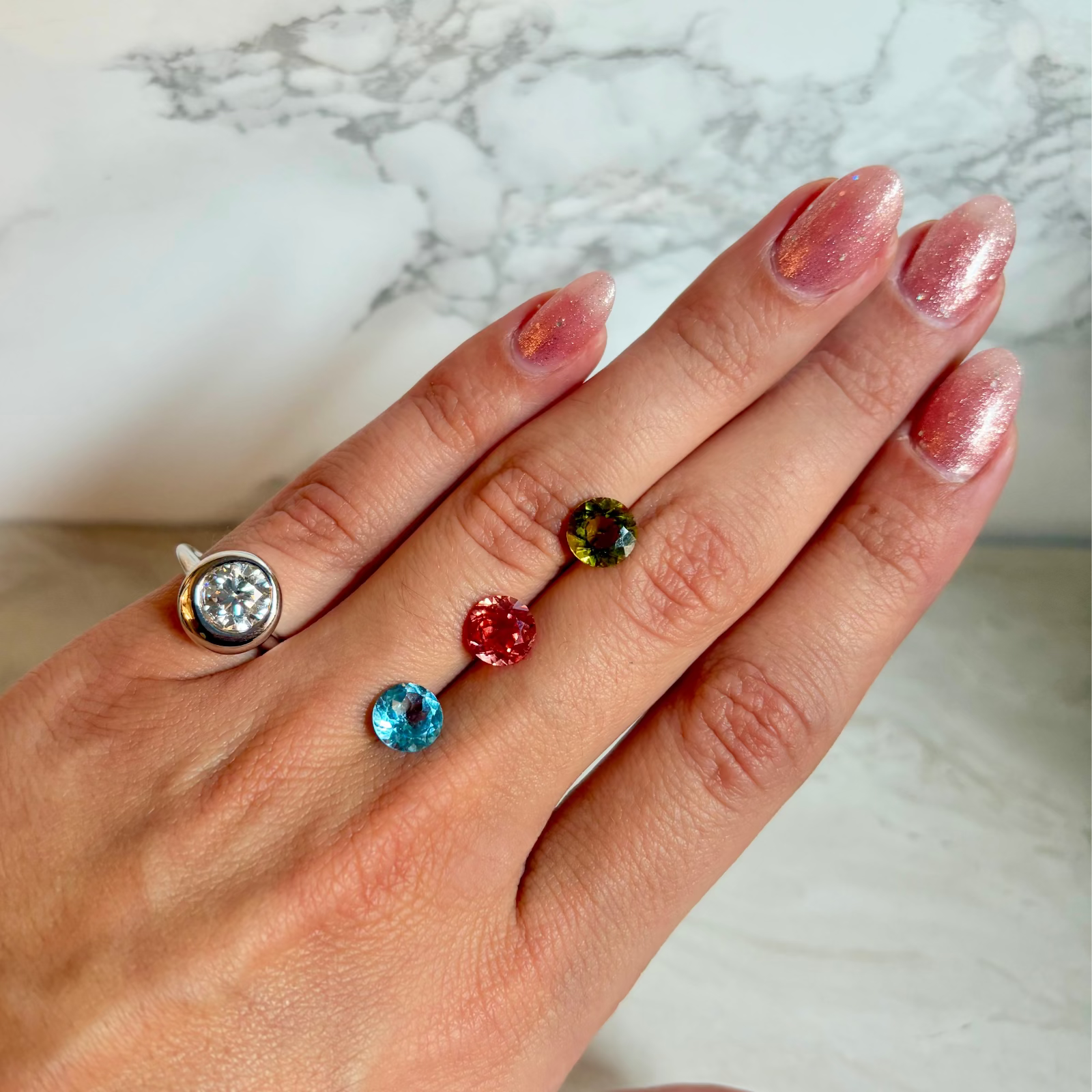 A hand with pink glitter nail polish wearing a white gold-toned ring with round diamond, and three small colorful gemstones (green, red, and blue) next to it.