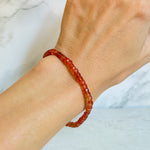 A close-up of a person's wrist wearing a beaded bracelet made of orange faceted padparadsha sapphire beads, set against a light marble background.