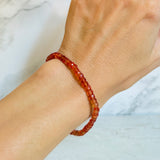 A close-up of a person's wrist wearing a beaded bracelet made of orange faceted padparadsha sapphire beads, set against a light marble background.