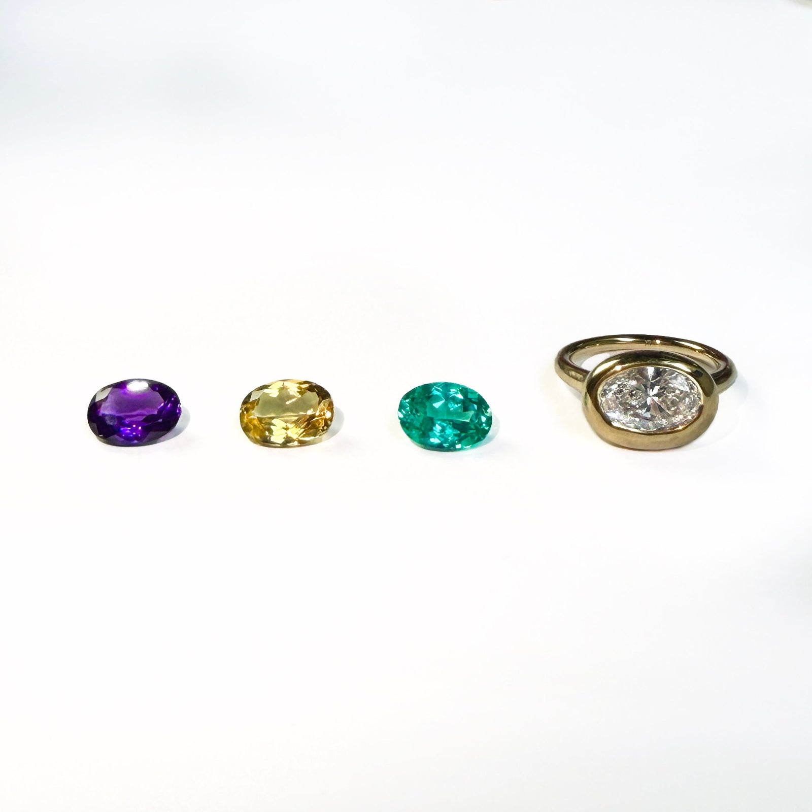3 colorful oval shaped gemstones next to a yellow gold molten solitaire oval-shaped ring on a white background.