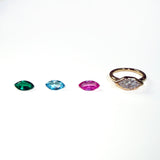 3 colorful marquise shaped gemstones next to a yellow gold molten solitaire marquise-shaped ring on a white background.