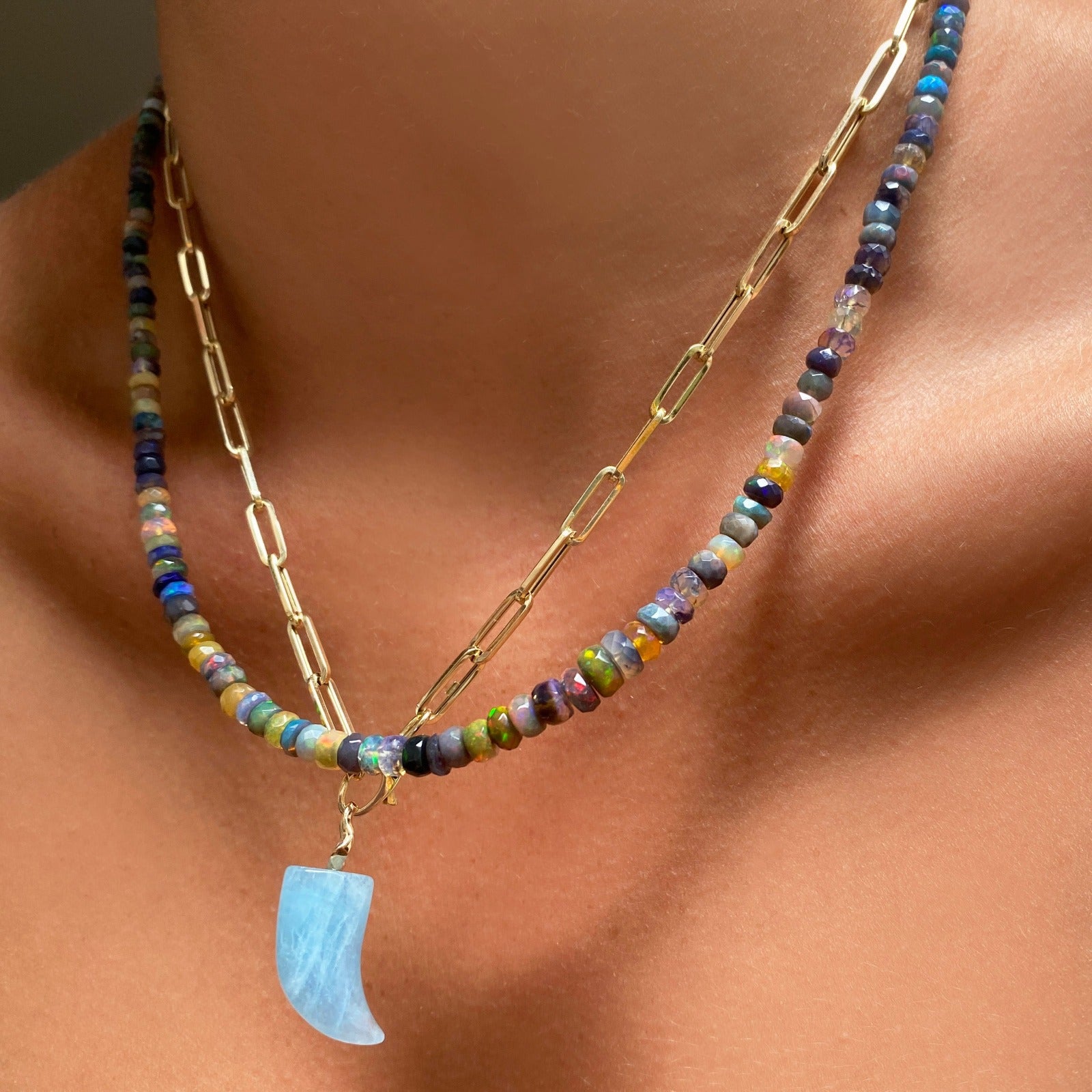 Shimmering beaded necklace made of faceted opals in shades of green, yellow, and blue on a gold linking ovals clasp. Styled on a neck layered with a chunky paperclip chain necklace and amazonite horn charm. 