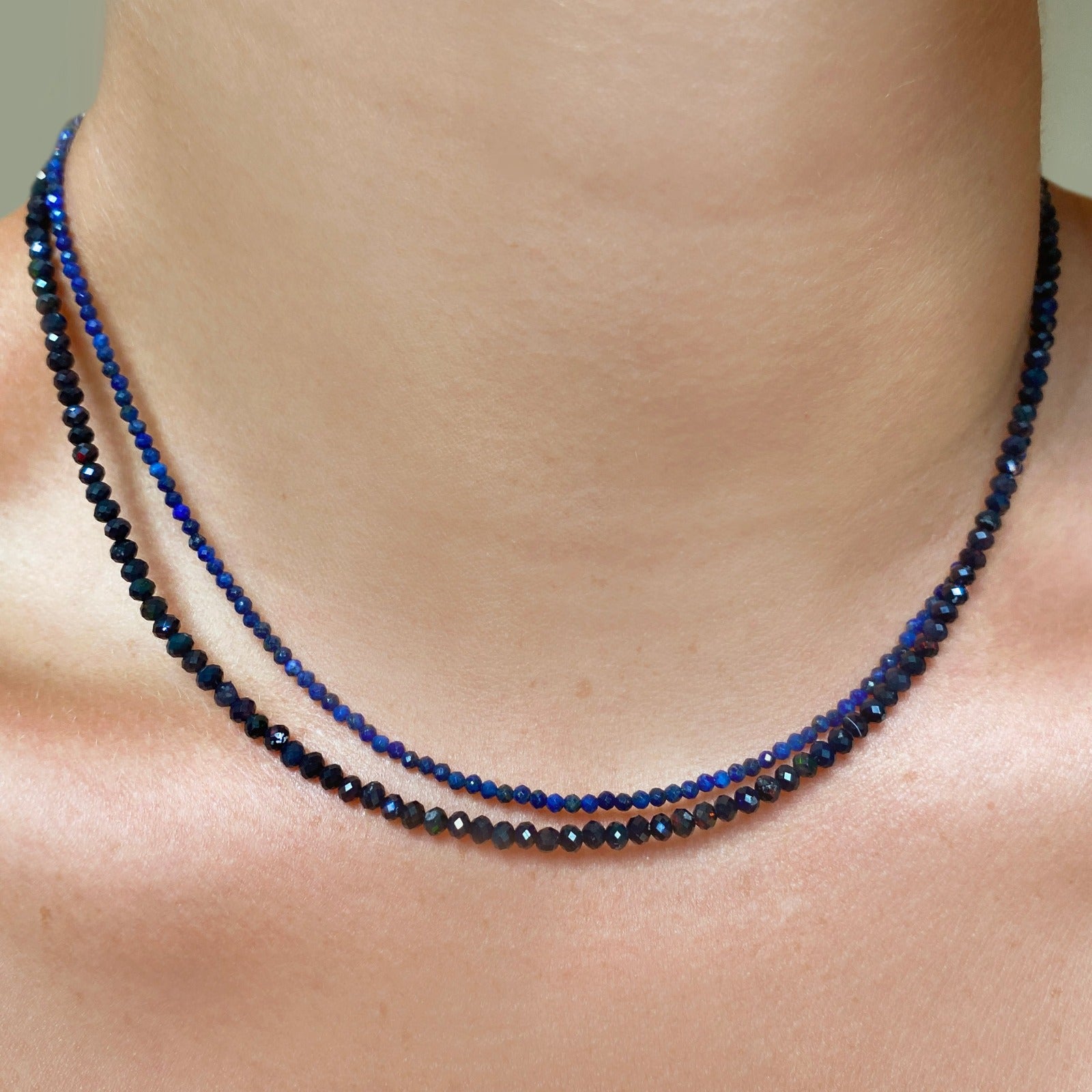 Shimmering beaded necklace made of 2.5mm faceted opals in shades of black on a gold linking lobster clasp. Styled with a navy blue 2.5mm faceted opal necklace on a gold linking ovals clasp.