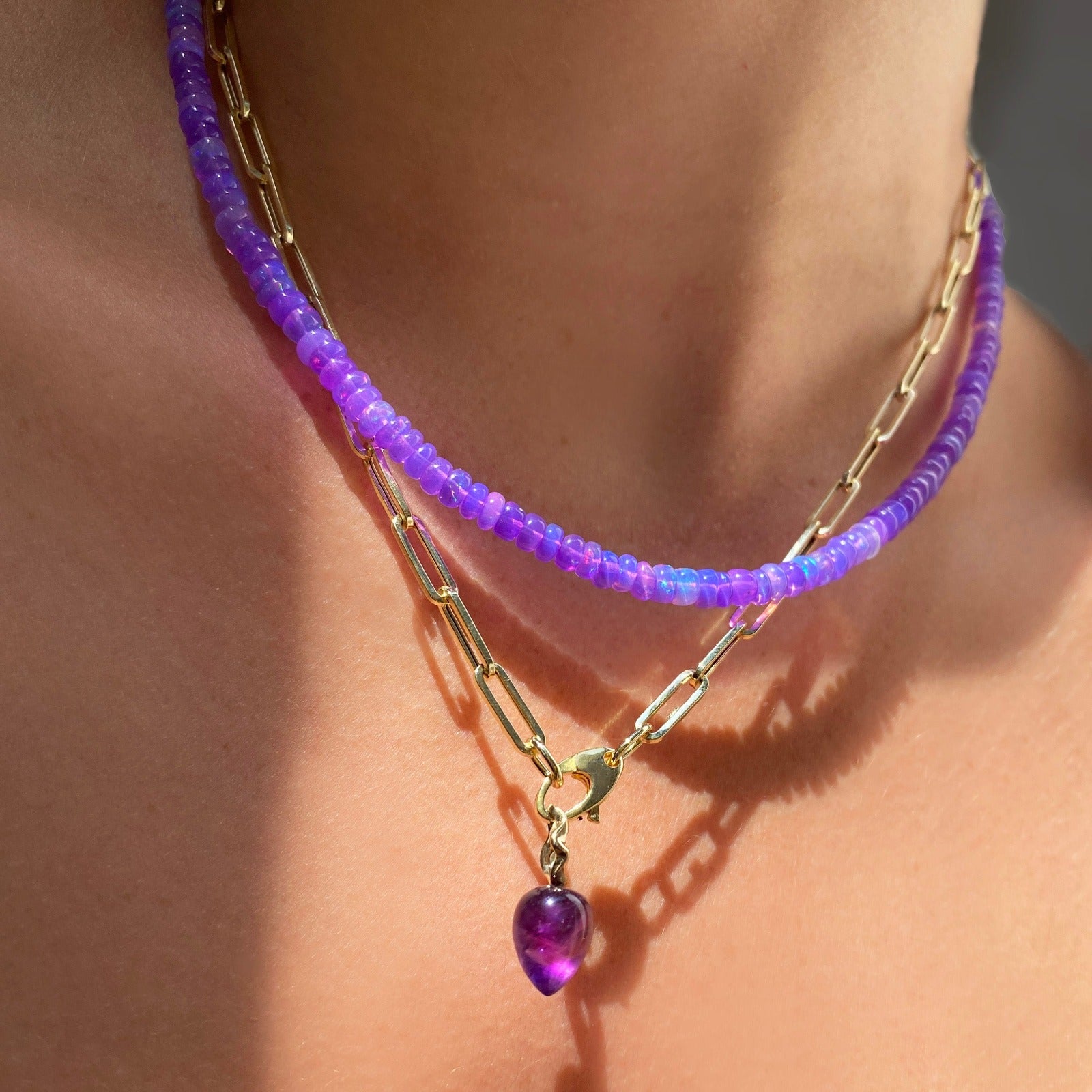 Shimmering beaded necklace made of smooth opal rondels in shades of violet on a slim gold oval clasp. Styled on a neck with with paperclip charm necklace and acorn drop charm.
