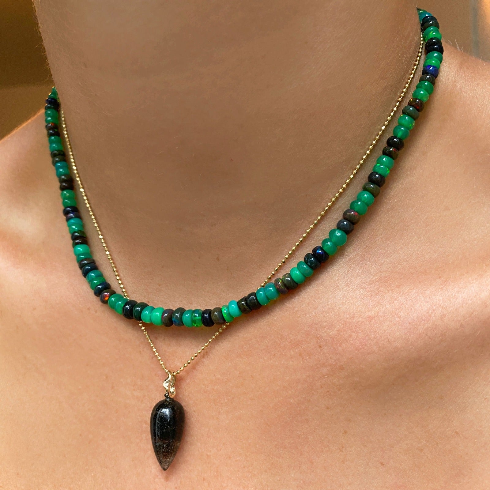 Shimmering beaded necklace made of smooth opal rondels in shades of green and black on a slim gold lobster clasp. Styled on a neck layered with a diamond cut bead chain necklace and acorn drop charm. 