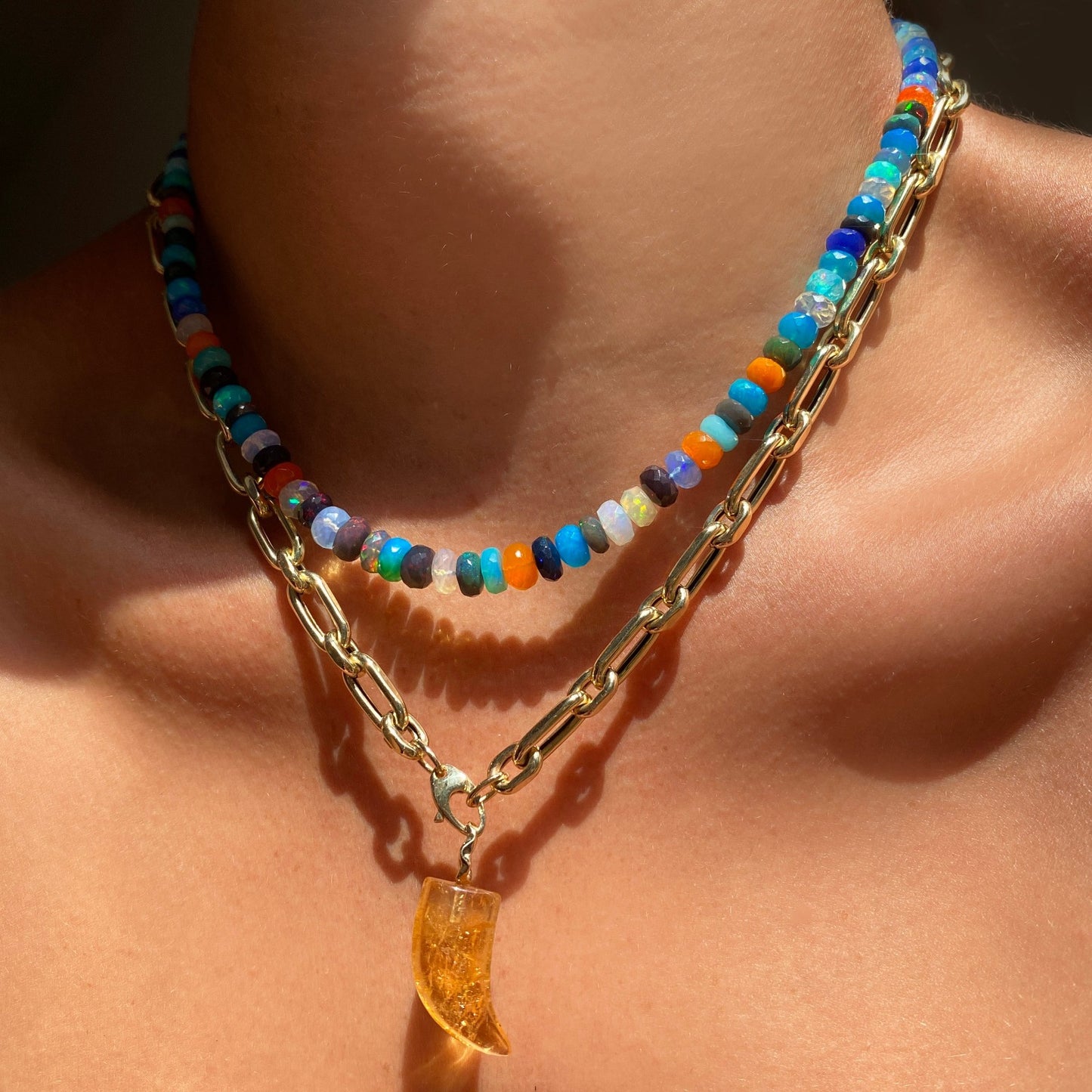 Shimmering beaded necklace made of faceted opals in shades of fiery blues, greens, teal, black, orange, and clear on a gold linking ovals clasp. Styled on a neck layered with a diamond cut link chain necklace and horn charm.