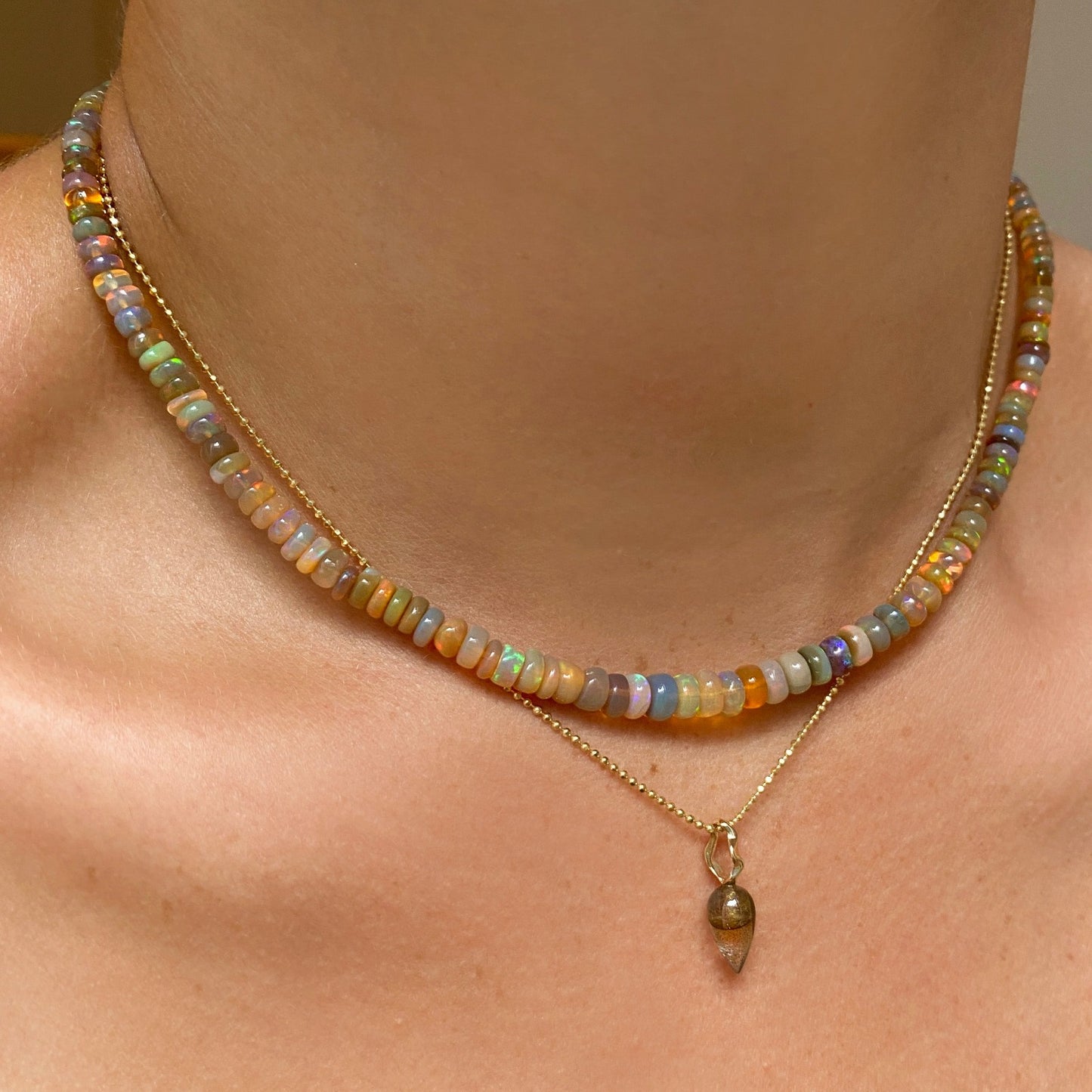 Shimmering beaded necklace made of smooth opal rondels in shades of light yellow, light orange, white, and brown on a slim gold lobster clasp. Styled on a neck layered with the bead chain necklace and acorn drop charm.