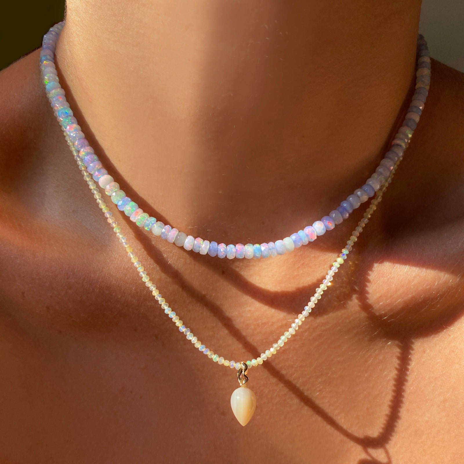 Shimmering beaded necklace made of faceted opals in shades of light pastel blue and white on a gold linking ovals clasp. Styled on a neck layered with an acorn drop charm.