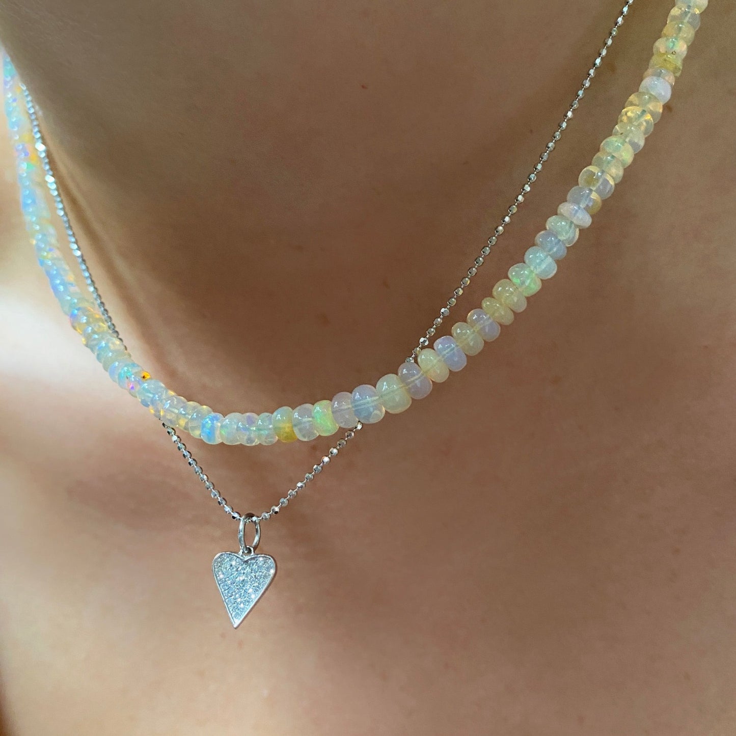 Shimmering beaded necklace made of smooth opal rondels in shades of clear natural opals on a slim gold lobster clasp. Styled on a neck layered with a diamond cut bead chain necklace and medium pave heart charm. 