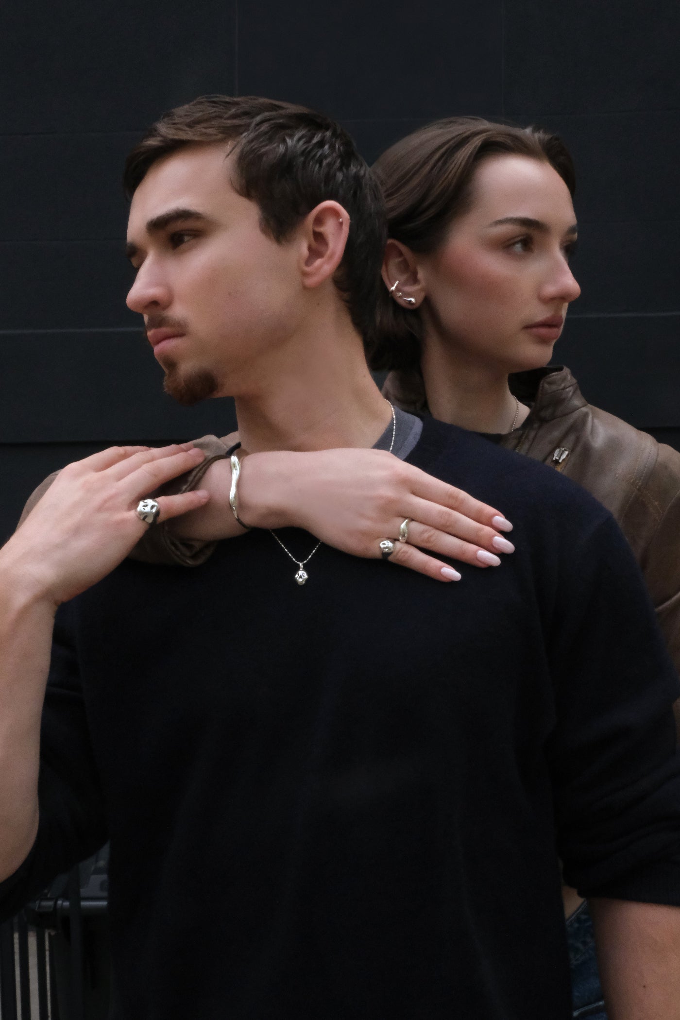 Two people standing close together with one person's hand on the other's shoulder, both wearing AMANDA PEARL molten sterling silver jewelry, against a dark background.