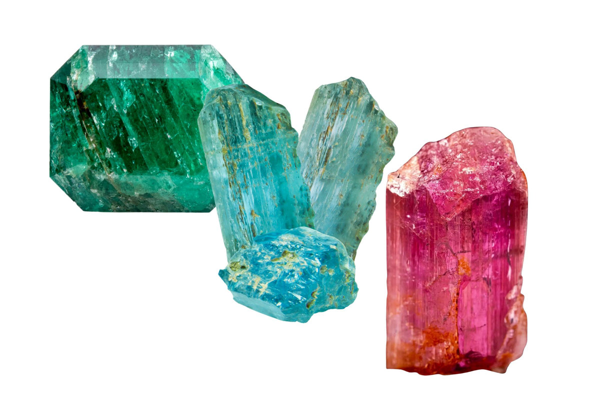 Rough, uncut emerald, blue topaz, and pink topaz stones sit grouped together on a white background.