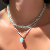 An AMANDA PEARL faceted aquamarine necklace layered over a White Zircon slim gemstone necklace with aquamarine drop charm with white gold ripple bale on a model's neck.
