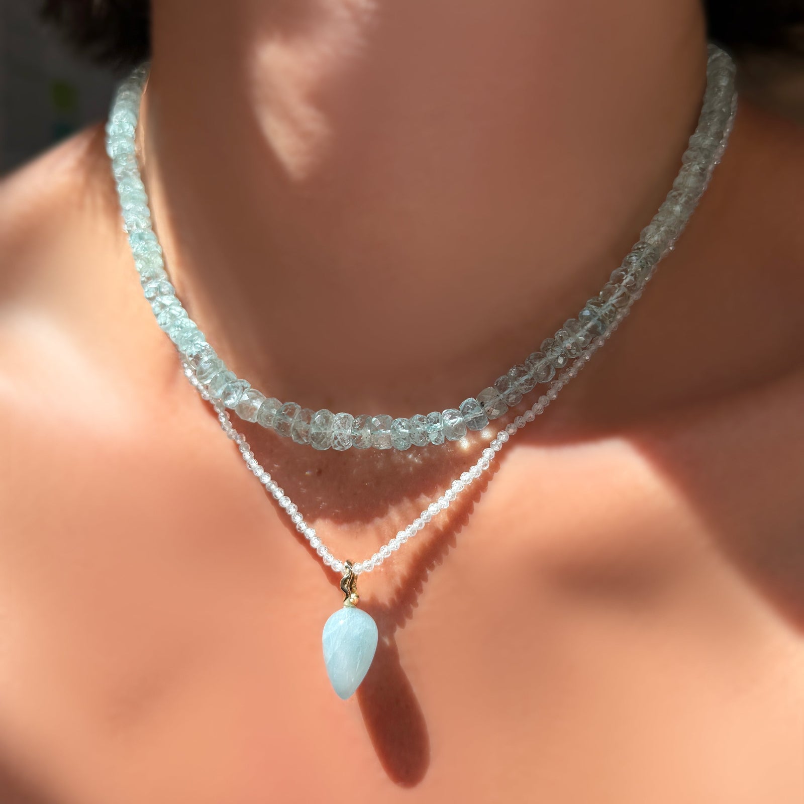 An AMANDA PEARL faceted aquamarine necklace layered over a White Zircon slim gemstone necklace with aquamarine drop charm with white gold ripple bale on a model's neck.