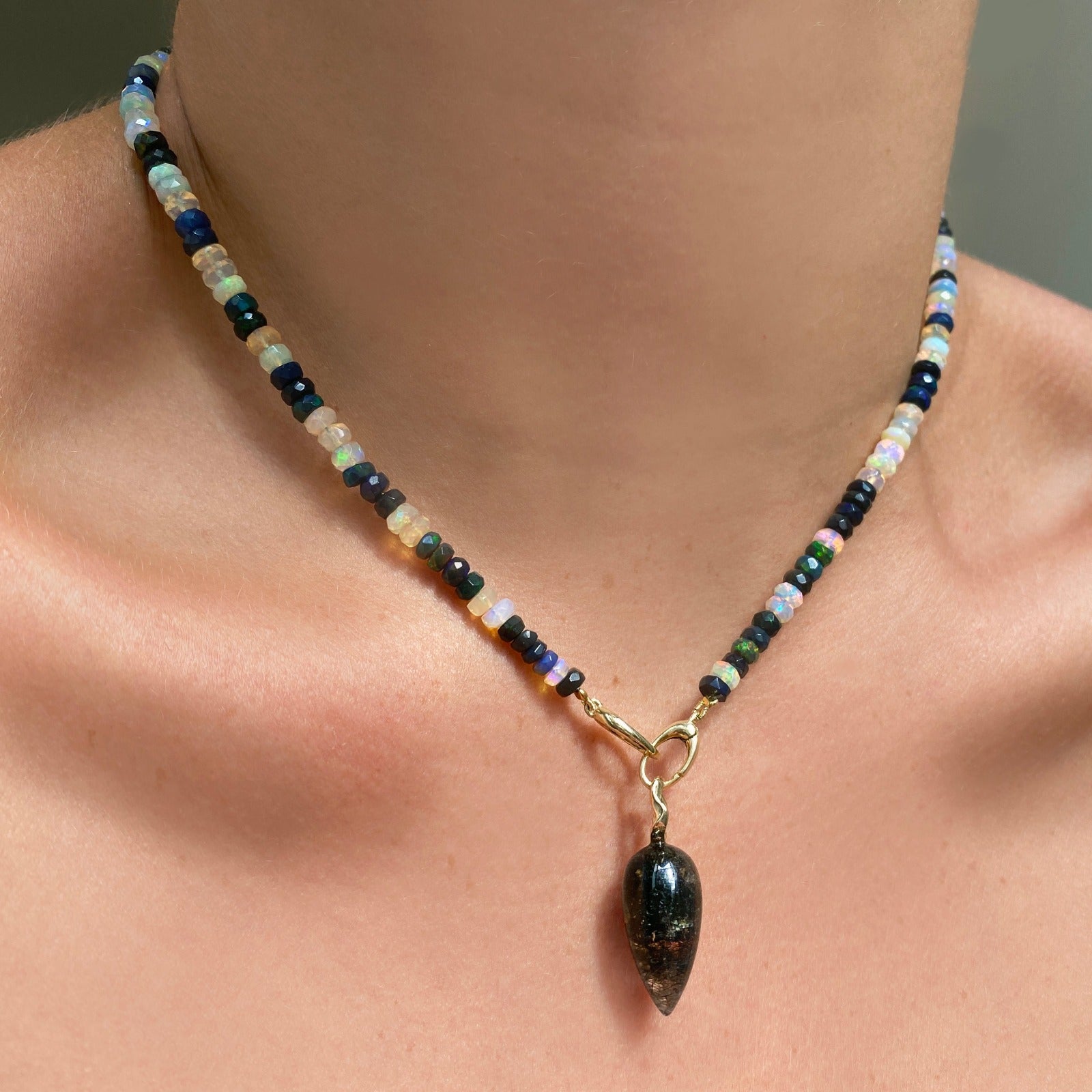 Shimmering beaded necklace made of faceted opals in shades of fiery pastels, grey, teal, and black on a gold linking ovals clasp. Styled on a neck layered with an acorn drop charm.
