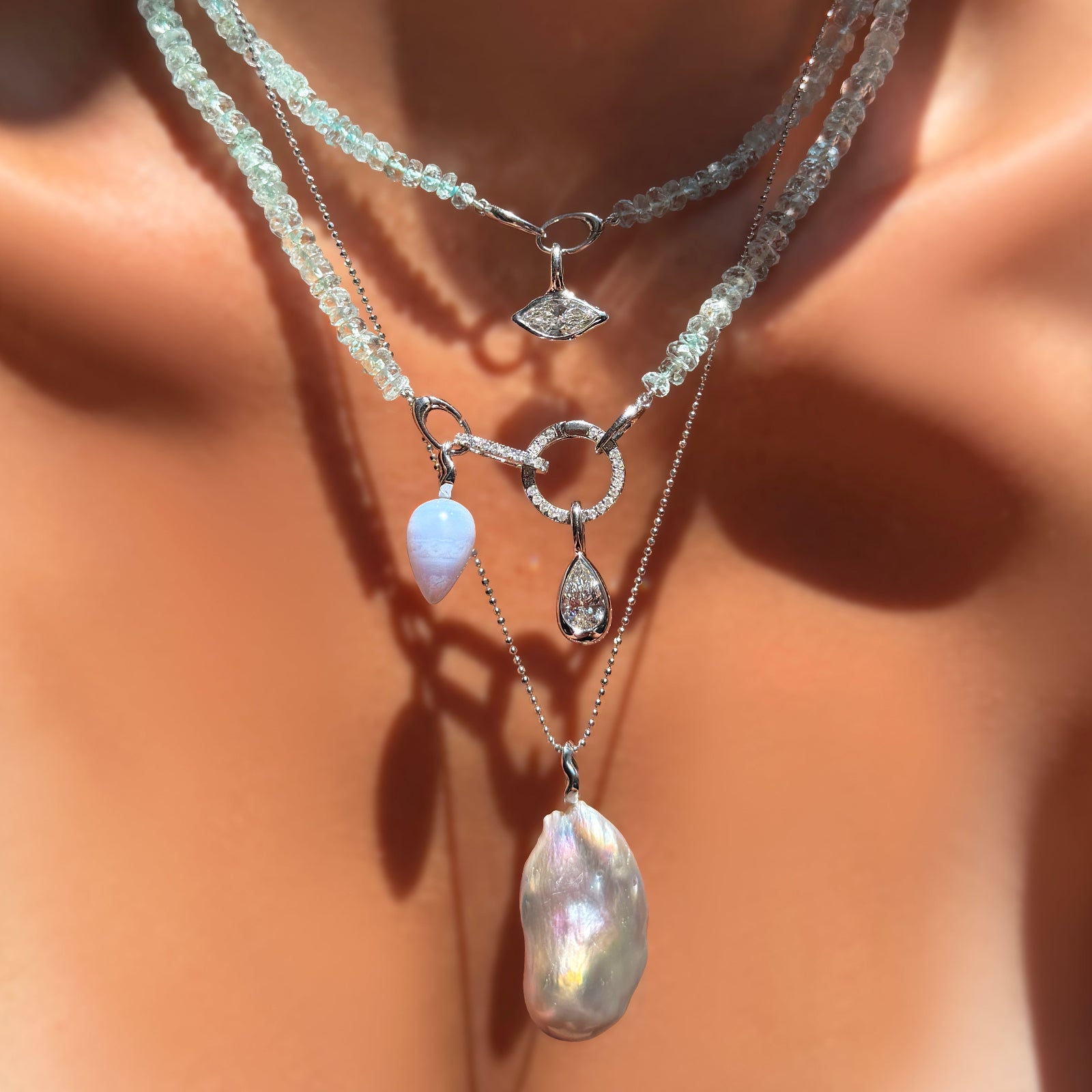 Faceted Aquamarine necklaces layered on a model's neck with molten solitaire charms with diamonds, diamond pavé charm locks, a blue lace agate drop charm  and baroque pearl charm on recycled white gold ripple bales and adiamond cut bead chain.
