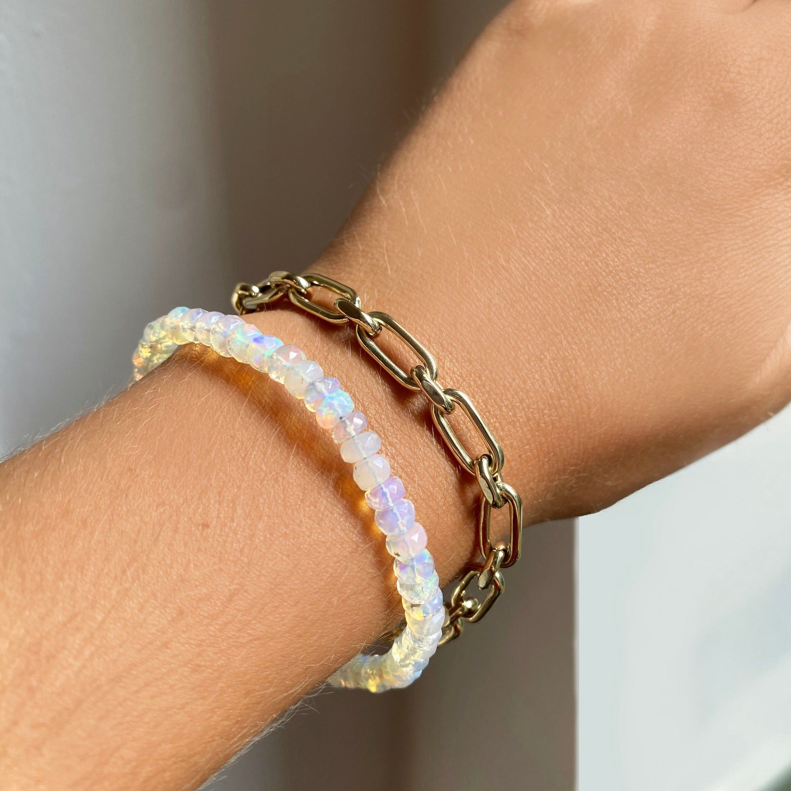 Shimmering beaded bracelet made of faceted opals in shades of milky white and clear on a gold linking lobster clasp. Styled on a wrist layered with diamond cut link bracelet.