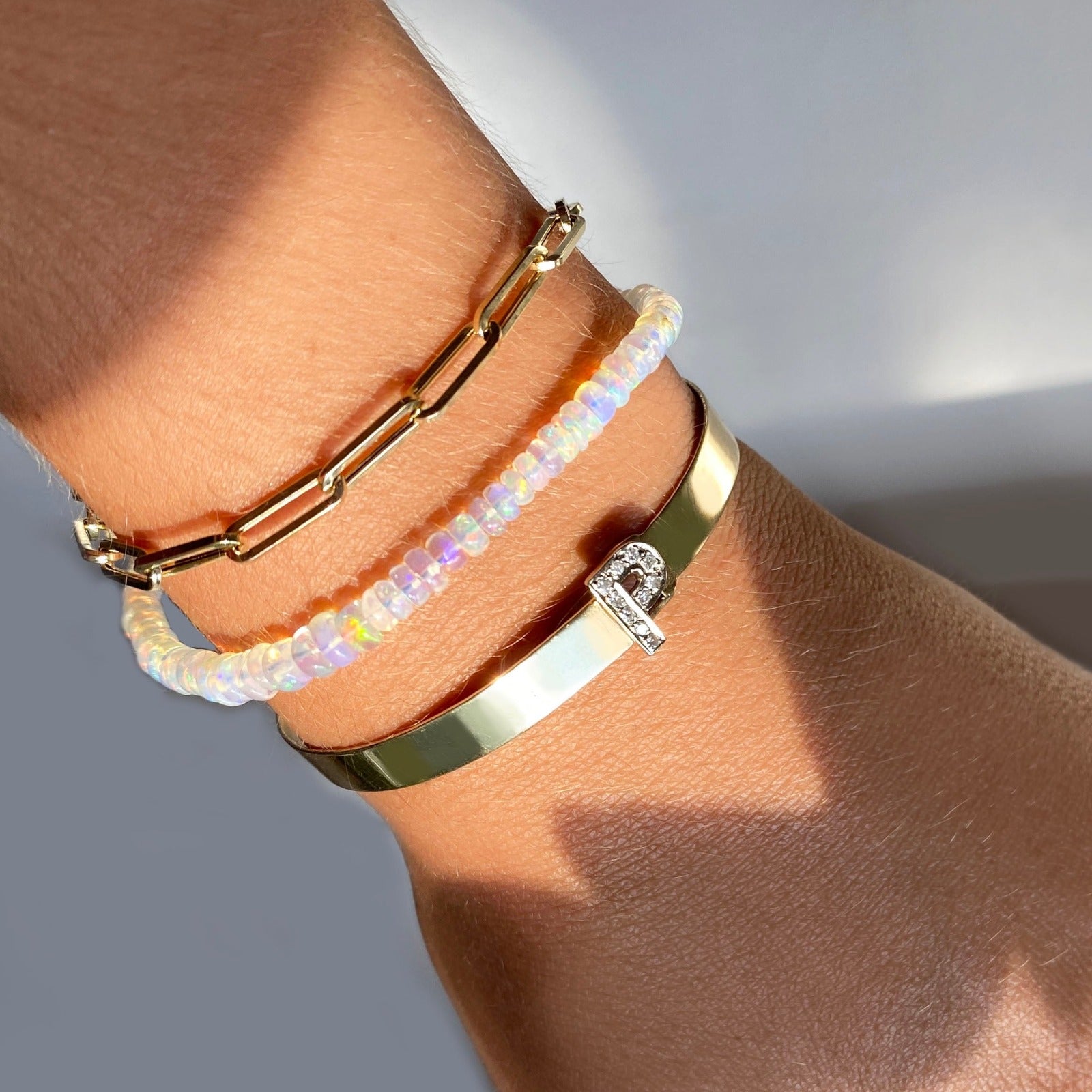 Shimmering beaded bracelet made of faceted opals in shades of milky white and clear on a gold linking lobster clasp. Styled on a wrist layered with a chunky paperclip chain bracelet and diamond letter bangle.