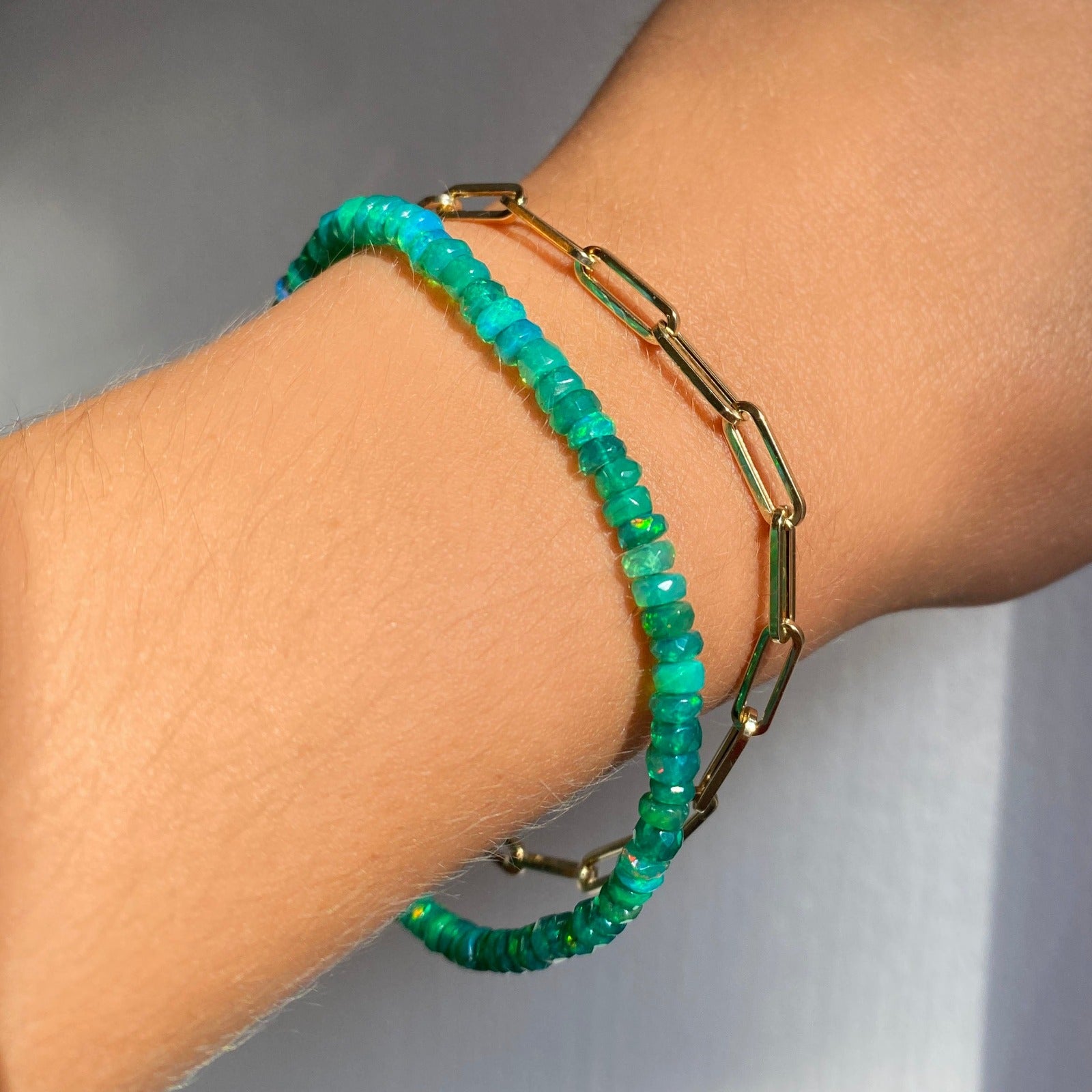 Shimmering beaded bracelet made of faceted opals in shades of green on a gold linking lobster clasp. Styled on a wrist layered with chunky paperclip chain bracelet.