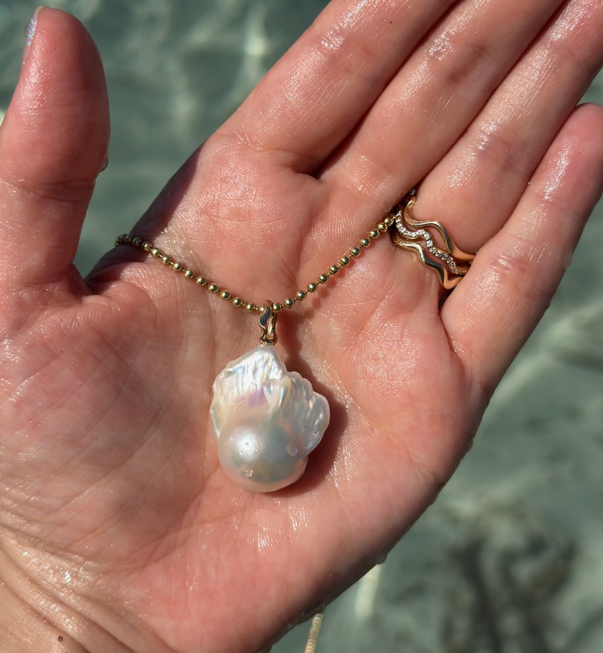 Hand holding a baroque pearl  charm on bead chain necklace with a blurred water background