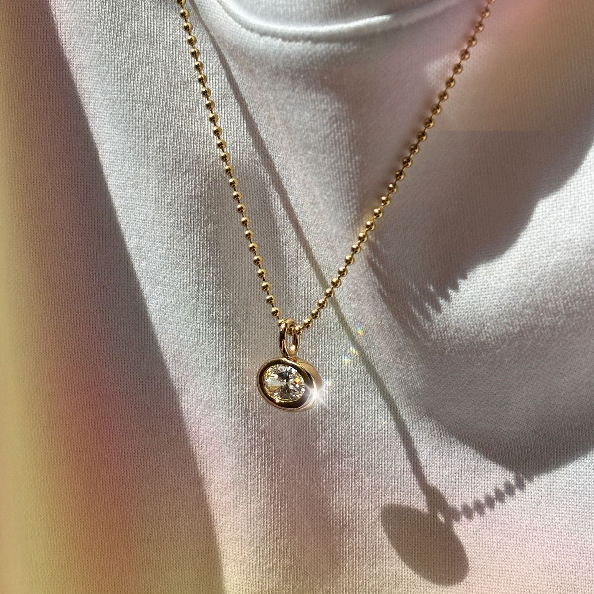14k gold molten solitaire charm with oval diamond hanging from a bead chain in sunlight.