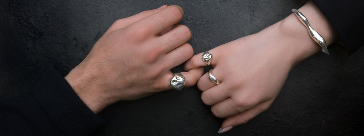 A couple locks pinkies against a black background. The man wears a skull pinky ring, as does the woman. She also wears a molten sterling ring and molten cuff bracelet, with rounded smooth lines. 