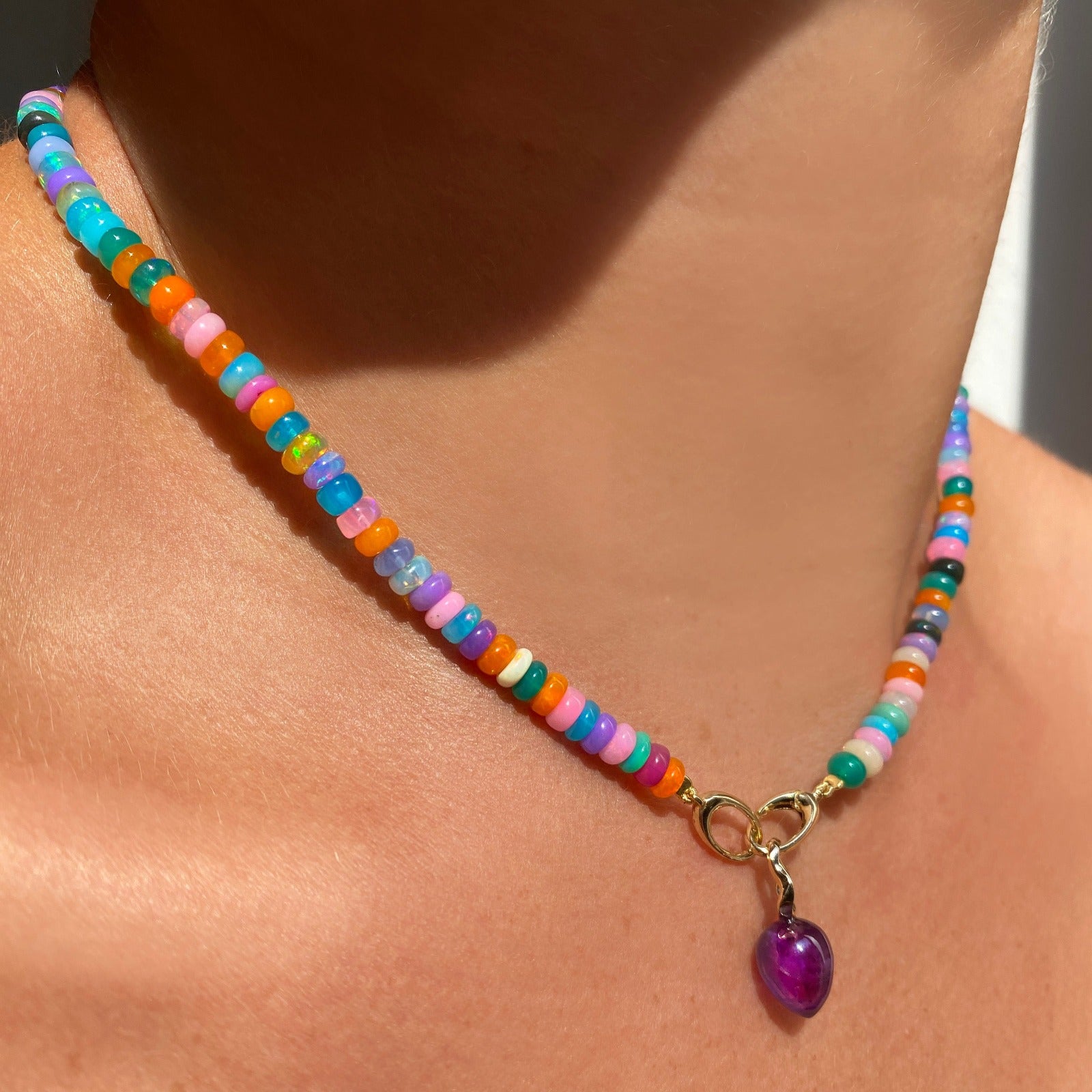 Shimmering beaded necklace made of smooth opal rondels in shades of fiery blues, hot pink, teal, and orange on a slim gold oval clasp. Styled on a neck layered with an acorn drop charm.