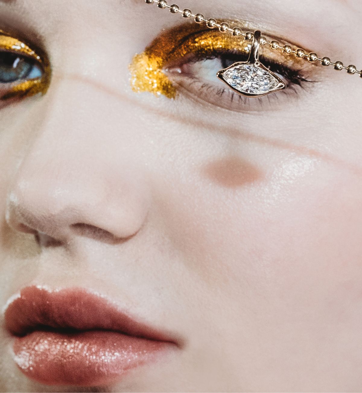 Molten solitaire charm on bead chain in front of a model's eye with gold eye shadow.