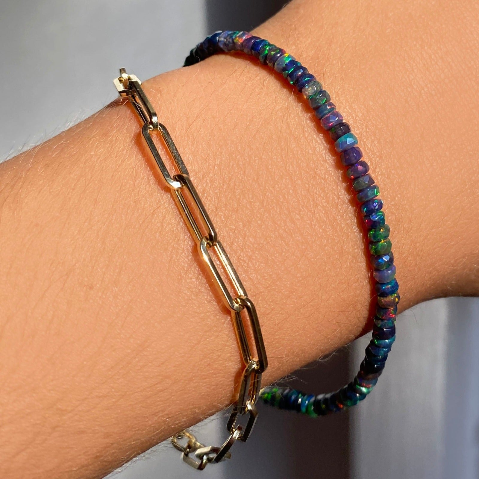 Shimmering beaded bracelet made of faceted opals in shades of black, blue, and green on a gold linking lobster clasp. Styled on a wrist layered with a chunky paperclip chain bracelet.
