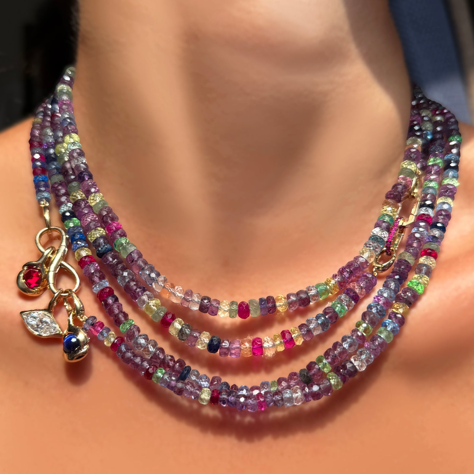 4 strands of night fever faceted sapphires with a molten infinity charm lock holding molten knot charms with star sapphire and rose cut ruby, along with a marquise molten solitaire charm, and two ruby charm locks on a model's neck.