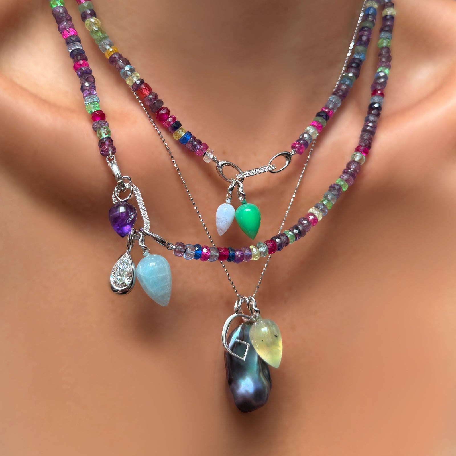 2 strands of night fever faceted sapphires with diamond pavé charm locks, colorful gemstone drop charms, a molten solitaire pear cut diamon charm, and a storm blue baroque pearl with letter G charm on a diamond cut bead chain on a model's neck.