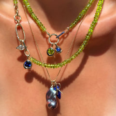 Two peridot gemstone necklaces with diamond and sapphire charm locks and diamond, sapphire, and peridot charms over a blue baroque pearl charm, Letter R charm, and lapis charm on a diamond-cut bead chain on a model's neck.