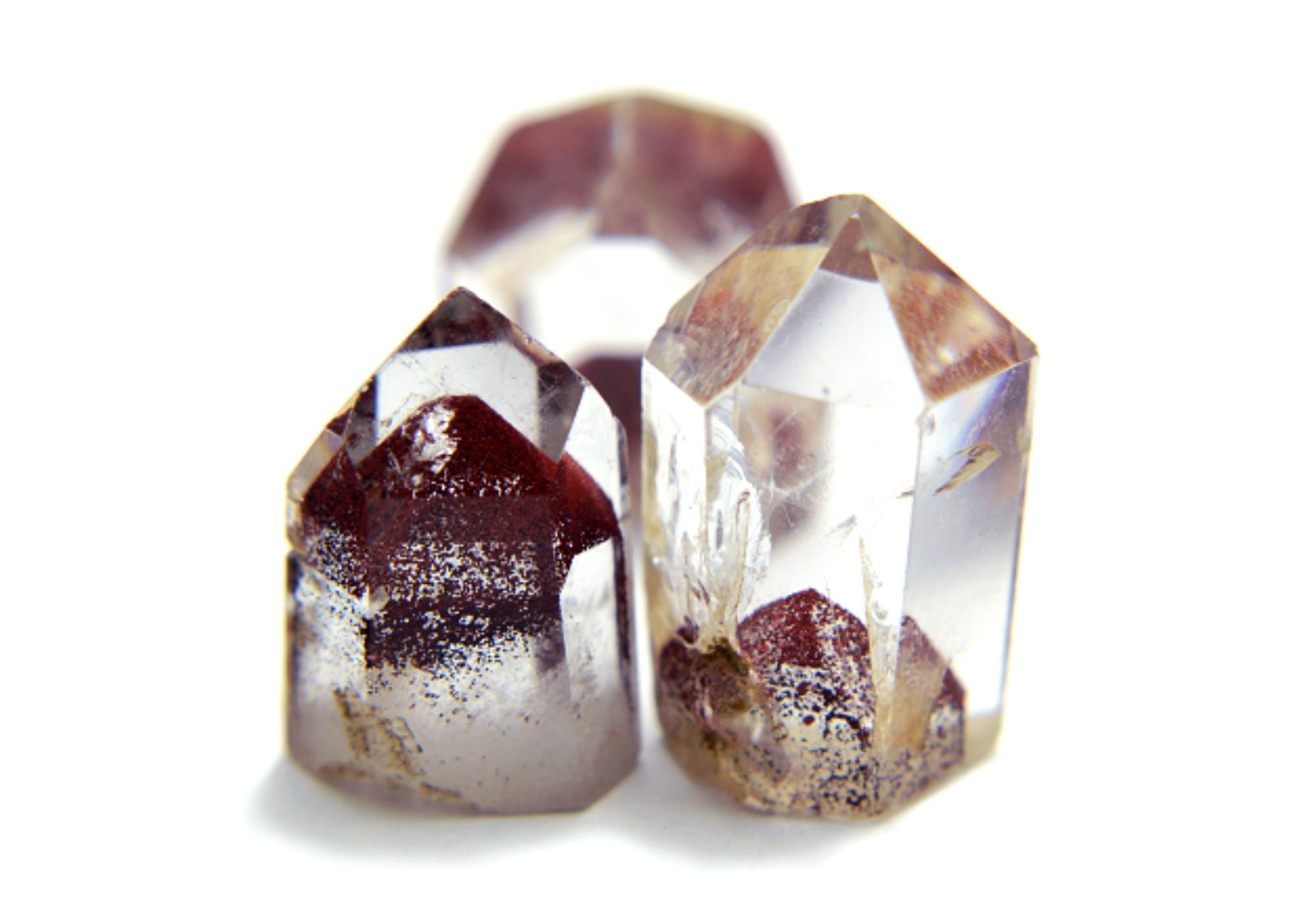 Three phantom clear quartz crystal points with embedded dark reddish-brown and metallic inclusions, photographed on a white background.