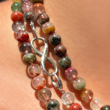 A close-up of a multiple strands of phantom quartz beaded necklaces worn on a person's neck. The necklace features polished round beads in a multicolor mix of rust, green, orange, yellow, red, and white, with a sterling silver evolution clasp holding a sterling silver infinity charm lock.
