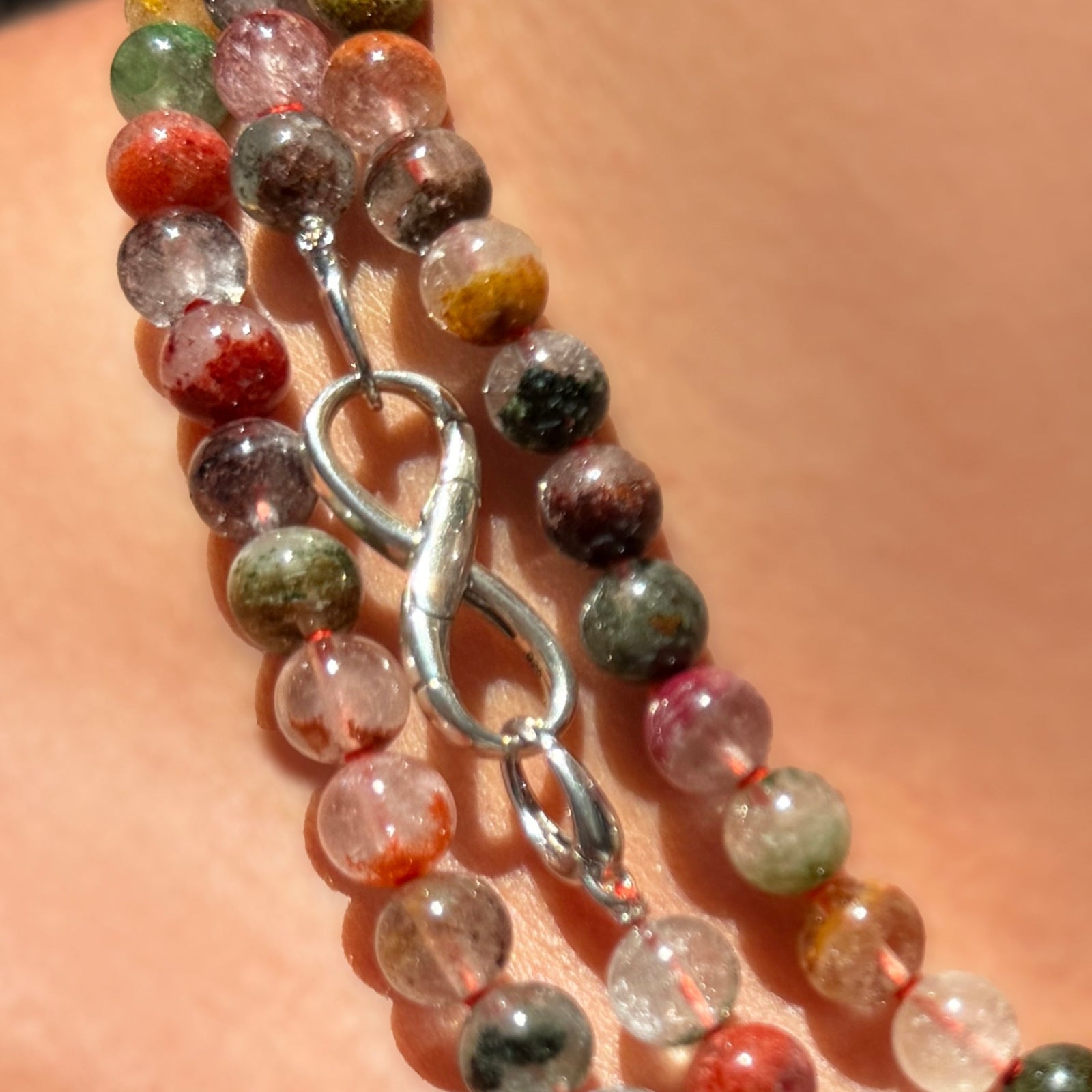 A close-up of a multiple strands of phantom quartz beaded necklaces worn on a person's neck. The necklace features polished round beads in a multicolor mix of rust, green, orange, yellow, red, and white, with a sterling silver evolution clasp holding a sterling silver infinity charm lock.