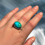An AMANDA PEARL recycled sterling silver ring with an oval turquoise cabochon worn on a model's finger, shown against a blurred blue-gray background.