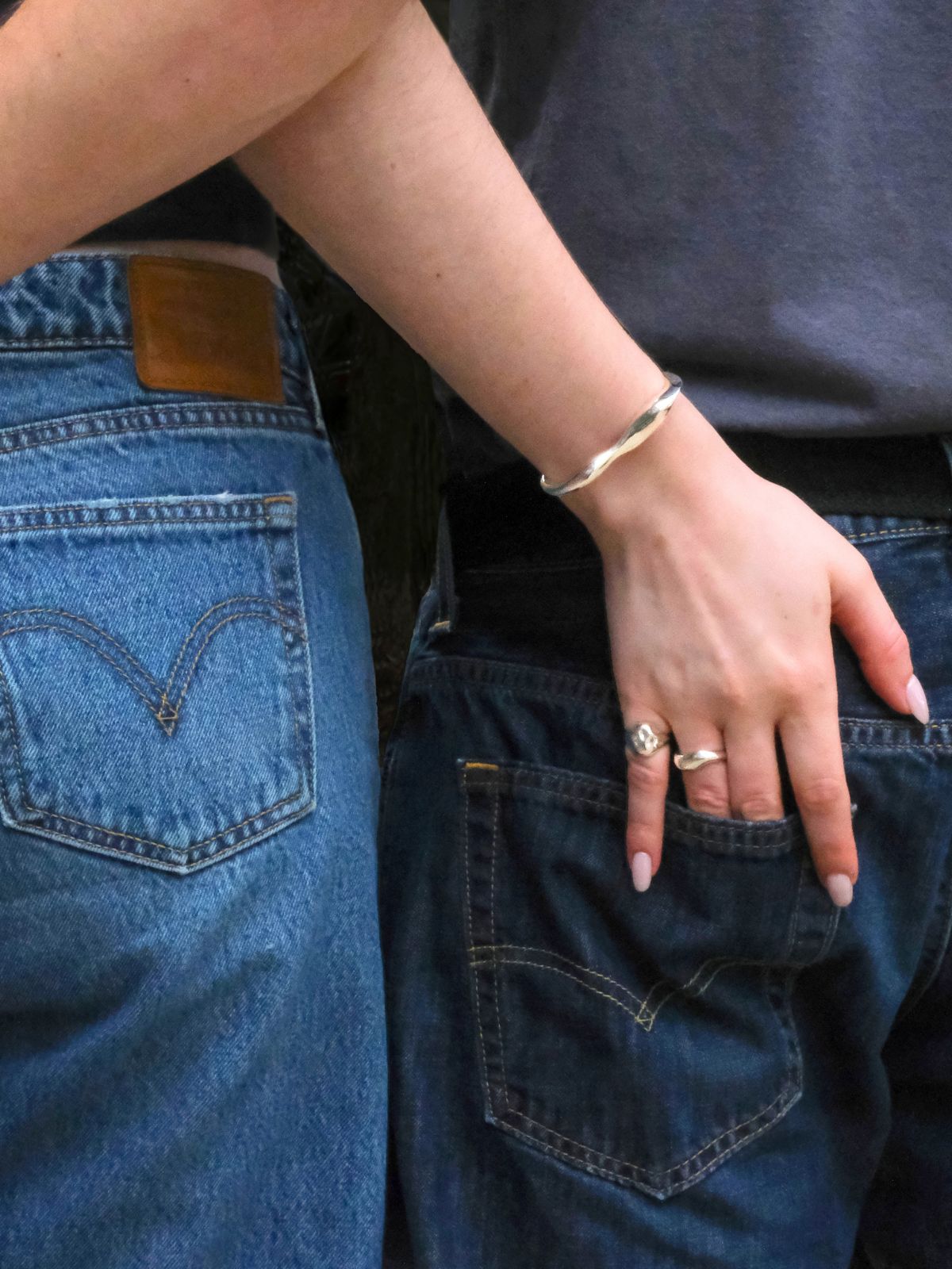 A close-up of a couple shot from behind, in jeans and black t-shirts. The woman's hand descends into the man's back pocket. It shows off her AMANDA PEARL jewelry in recycled sterling silver. 
